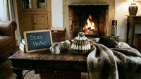 Cozy living room with fireplace, knitted hat, mugs, fur blanket, and chalkboard sign saying Stay Home in warm hygge interior.
