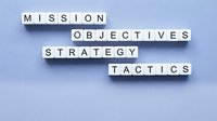 Letter cubes spelling mission, objectives, strategy, and tactics arranged in crossword style on a light blue background for business planning.