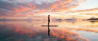 Stand-up paddleboard athlete balancing on board during golden hour sunrise or sunset over calm water with dramatic sky reflections.
