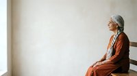 Elderly woman with gray hair wearing rust-colored clothing and headscarf sits alone by window in contemplative pose, suitable for senior lifestyle, wellness, and emotional storytelling content.