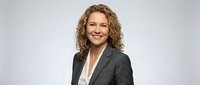 Professional middle-aged Caucasian businesswoman with curly blonde hair smiling confidently in dark suit jacket on neutral background for corporate use.