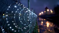 Razor wire security fence illuminated with decorative white LED string lights at dusk creates contrast between harsh barrier and soft glowing ambiance.