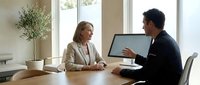 Professional business meeting with mature Caucasian businesswoman and young Asian businessman discussing project at modern office conference table.