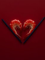 Two cooked shrimp arranged in heart shape on dark red background with dramatic lighting and shadows, creating romantic seafood concept for Valentine's Day menus and promotions.