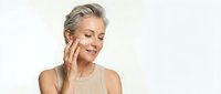 Mature blonde woman applying skincare cream with gentle touch, eyes closed in peaceful expression. Anti-aging beauty routine and facial care concept.