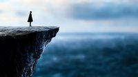 Silhouette of lone woman standing on cliff edge against moody blue ocean and cloudy sky background, representing solitude, courage and life challenges.