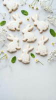 Easter bunny cookies with white icing and sprinkles arranged on marble surface with chamomile flowers and mint leaves for spring holiday baking celebration.