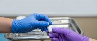 Healthcare worker in blue gloves passing small medical vial to colleague in purple gloves in clinical laboratory setting for pharmaceutical research.