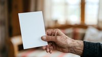Senior man with wrinkled hands holding blank white card indoors, suitable for greeting cards, invitations, announcements, and promotional messaging designs.