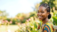 Young African woman with braided hairstyle wearing colorful traditional patterned clothing, smiling outdoors in sunny garden setting with blooming flowers and green foliage background.