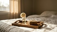 Wooden breakfast tray with coffee cup and flower on unmade bed in sunlit bedroom. Morning routine, hospitality service, cozy home interior concept.