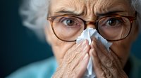 Senior Caucasian woman with glasses blowing nose into tissue, showing cold or flu symptoms against dark background, close-up portrait in natural lighting.