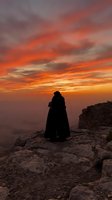 Silhouetted figure in dark cloak standing on rocky cliff edge gazing at dramatic orange and red sunset sky with golden clouds, contemplative mood for editorial and creative projects.