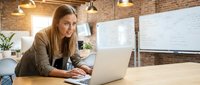 Young professional businesswoman working on laptop in modern office with brick walls and whiteboard for corporate presentations and meetings.