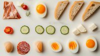 Fresh breakfast ingredients in rows including eggs, prosciutto, bread slices, cucumber, tomatoes and salami on light background, flat lay composition for menu design.