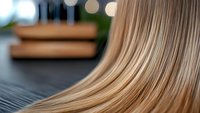Smooth flowing blonde hair strands on dark background with soft bokeh lights, close-up macro shot showing texture and shine of healthy straight hair.