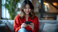 Young Asian woman in red blouse smiling while using smartphone on couch, expressing joy and engagement with mobile device in modern living room setting.