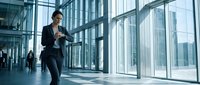 Professional businessman in dark suit checking smartphone while walking through modern glass office building corridor with city skyline view.