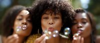 Three young African American women with natural curly hair blowing soap bubbles outdoors, expressing joy and playfulness in warm sunlight. Close-up portrait shows pure happiness.