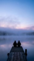 Two people sitting on wooden dock at peaceful lake during twilight hour with pastel sky colors reflecting in calm water for romantic getaway concepts.