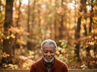 Senior man with gray beard and glasses wearing burgundy sweater smiling outdoors in autumn forest with golden foliage. Portrait for lifestyle and wellness content.
