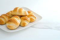 Fresh baked golden croissants served on white ceramic plate against marble background, soft natural lighting creates appetizing breakfast scene.