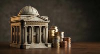 Classical bank building with dome and columns next to stacked golden coins on wooden surface representing financial institution wealth and monetary growth.