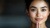 Close up portrait of young Asian woman with natural makeup and glowing skin touching chin thoughtfully against dark background, wellness and beauty concept.