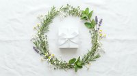 White gift box with ribbon centered in a round wreath of fresh herbs and wildflowers on white linen background.