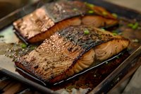Grilled salmon fillets with crispy skin and caramelized glaze on dark baking sheet, seasoned with herbs and spices, showing moist, flaky texture.