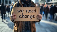 Young African American man holding cardboard sign with message during street protest, crowd in background, urban setting, social activism concept.