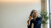 Mature woman with gray hair and South Asian ethnicity wearing dark blazer adjusting collar in modern office with plant, professional portrait for business and corporate use.