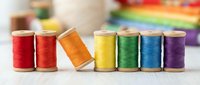 Colorful thread spools arranged in rainbow order on wooden surface for sewing, crafting, and textile projects with blurred fabric background.