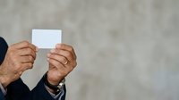 Mature man with brown skin holding blank white card in both hands against neutral beige wall background for messaging or advertising purposes.