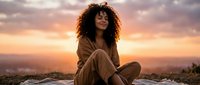 Young African American woman with curly hair meditating peacefully at sunset on mountain top, wearing cozy sweater, enjoying serene moment in nature.