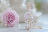 Elegant rose gold treble clef necklace pendant with crystal detail and delicate chain against soft pink rose background, romantic jewelry macro photography.