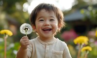 Joyful Asian toddler holding dandelion seed head in blooming garden, laughing with pure happiness among yellow flowers on sunny day.