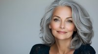 Confident mature woman with natural gray wavy hair and radiant smile wearing dark sweater against neutral background, representing beauty and aging gracefully.