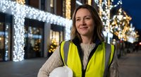 Female construction worker or safety inspector smiling at festive holiday street with Christmas lights and decorations in urban evening setting.