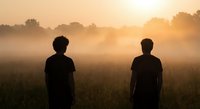 Two men silhouettes standing in misty field during golden sunrise with warm atmospheric lighting and peaceful countryside landscape.