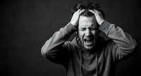 Stressed middle-aged Caucasian man holding head in hands screaming with anguish and frustration against dark background in black and white portrait.