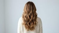 Young woman with long wavy blonde hair wearing white knit sweater from behind against neutral background for hair care and beauty concepts.