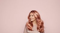Smiling woman with long wavy rose gold hair against pink background, suitable for beauty, cosmetics, and hair care marketing materials.