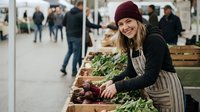 Young blonde woman vendor in burgundy beanie and apron smiling while arranging fresh vegetables at indoor farmers market stall with customers shopping.