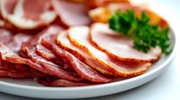 Sliced cured meat platter with prosciutto, salami, and ham garnished with fresh parsley on white ceramic plate, selective focus on meat texture.