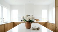 Bright minimalist kitchen with warm wood cabinets, white countertops, and a rustic ceramic vase of white peonies on the island.