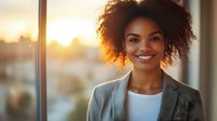 Young African American businesswoman with natural curly hair smiling confidently at sunset, wearing grey blazer and white top against urban background with warm lighting.