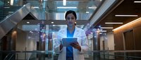 Young Asian male doctor in white coat using digital tablet with holographic medical interface in modern hospital corridor for healthcare technology.