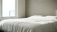 Unmade bed with white linens in minimalist bedroom with natural light from window and neutral textured walls, suitable for interior design and lifestyle content.