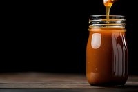 Sweet golden caramel sauce pouring into glass mason jar on dark wooden background, dramatic food photography with copy space for dessert recipes.